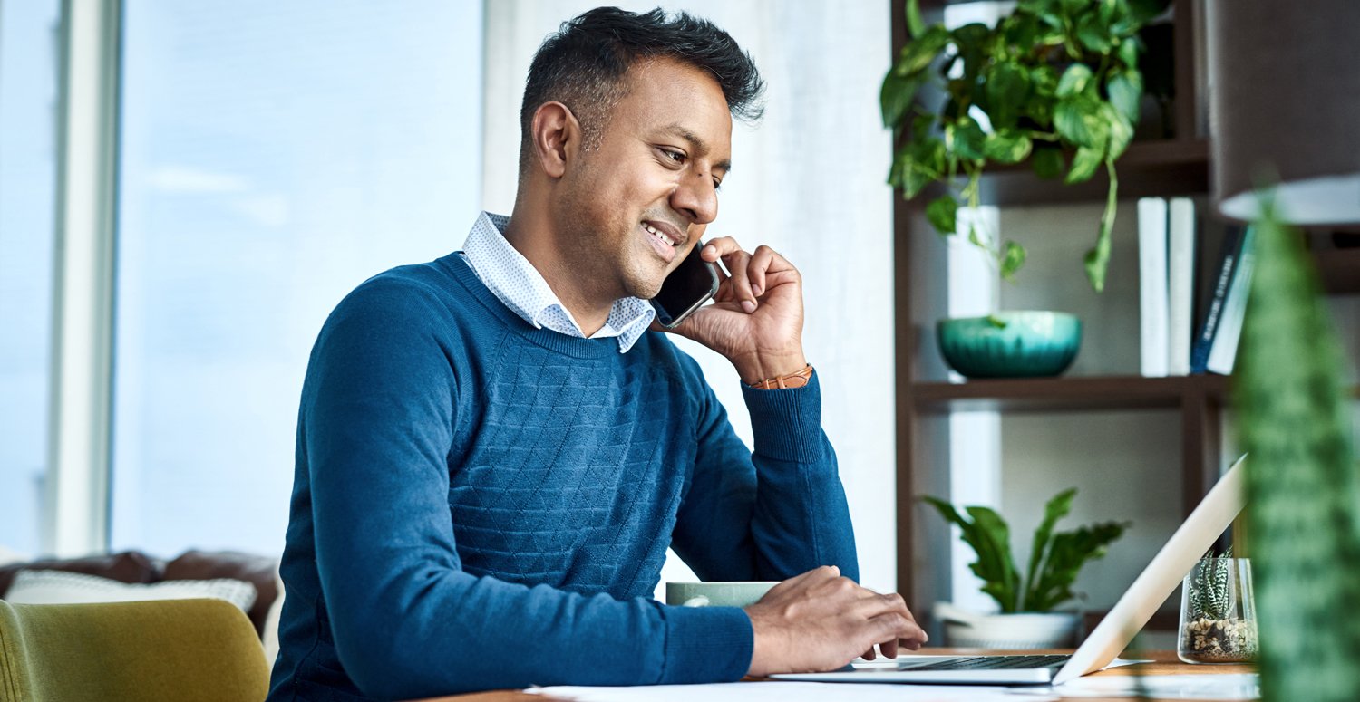 Man wearing a blue sweater smiling with his cellphone in his left hand and his right hand using his laptop. 