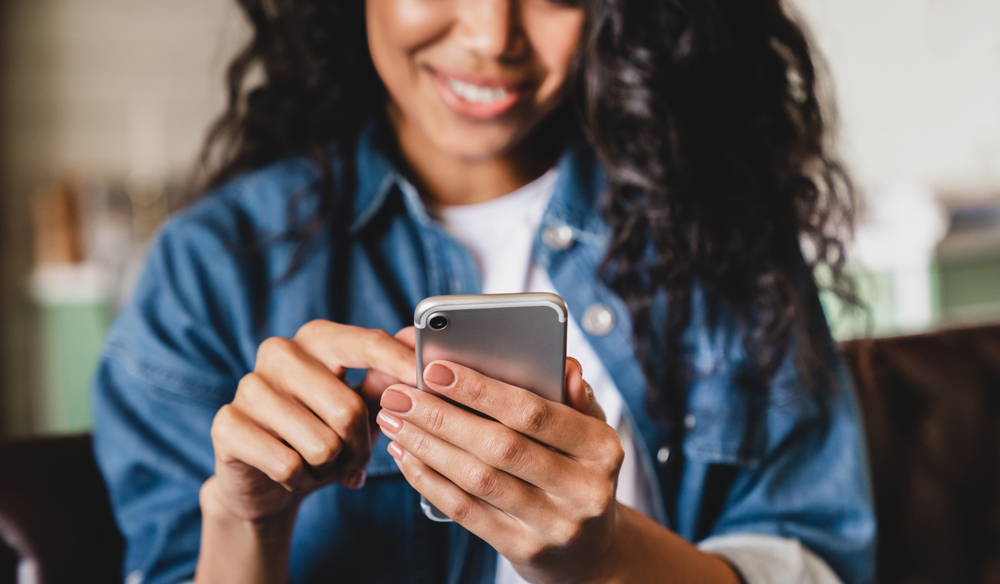 Woman wearing a denim jacket over a white shirt smiling and typing on her phone.