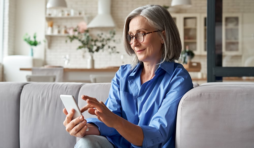 Woman sitting on a couch wearing a blue collared shirt and glasses clicking on her phone.