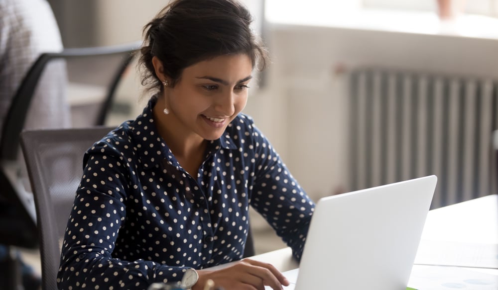 Woman sitting down wearing a blue and white polka dot shirt looking at a laptop and smiling.