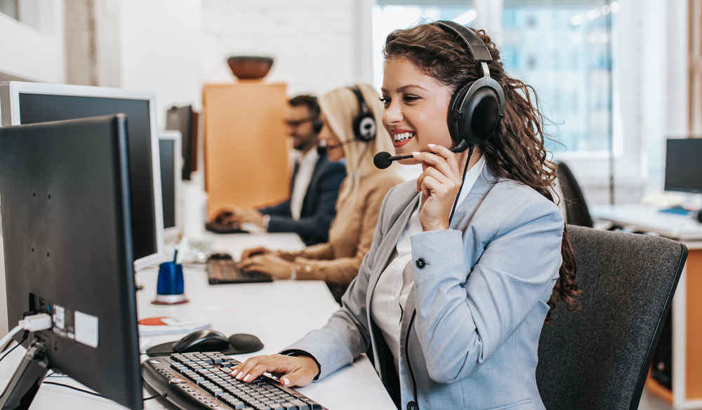 Woman wearing a grey blazer smiling while looking at her computer with a headset on.