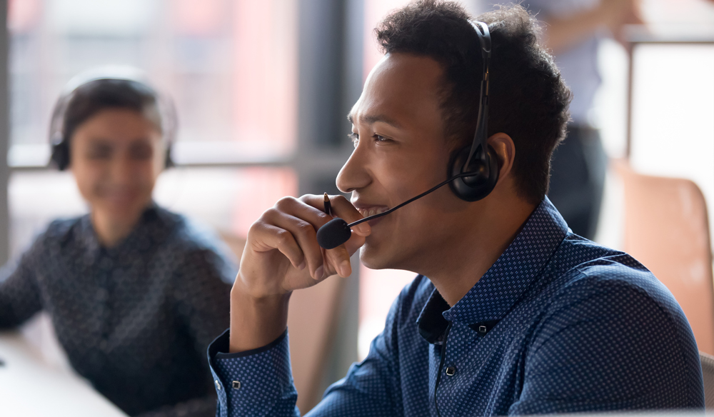 Man smiling wearing a blue collared shirt smiling and speaking into a headset.