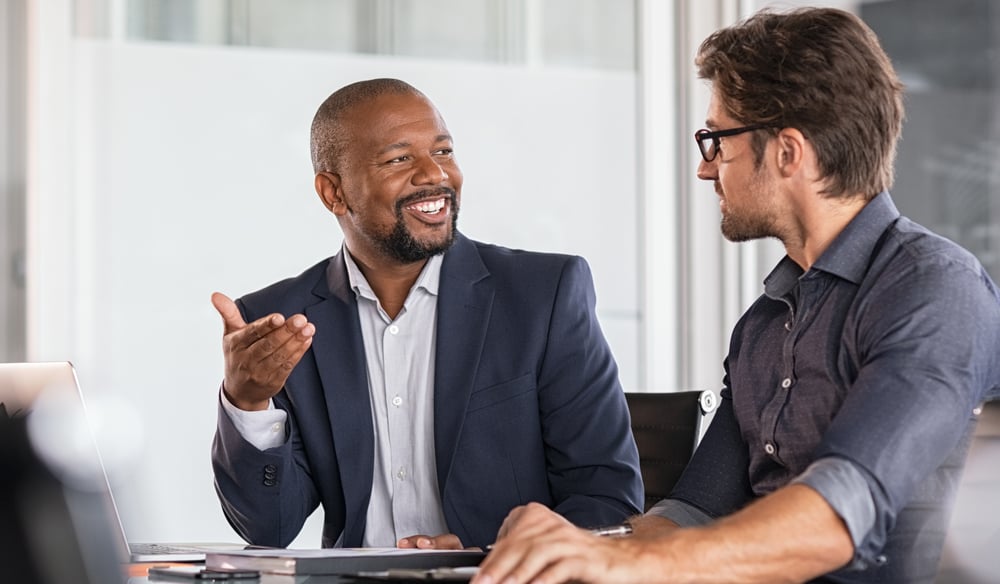 Two males sitting down in an office speaking to each other.